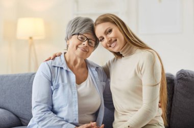 Gülümseyen yaşlılar ve genç kadınlar rahat koltukta oturur, kucaklaşır ve kameraya bakarlar. Mutlu kıdemli anne ve yetişkin kızı kucaklaşıyorlar, sevgi, bağ ve sıcaklığı iki nesil arasında paylaşıyorlar..