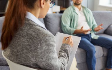 Stresli bir adam terapi seansı sırasında elleri bağlı bir şekilde koltukta oturur. Bir kadın psikolog dinlerken not alır. Sahnede danışmanlık, destek, akıl sağlığı ve duygusal bakım gösteriliyor..