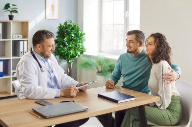 Doktor ve mutlu çift hastane ofisinde konsültasyonda. Genç eş ve koca, aile planlaması kliniğindeki doktordan iyi haberler almak için birlikte oturuyorlar. Sağlık, hamilelik, tüp bebek konsepti.