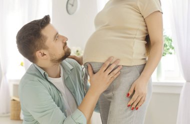 Hamile karısının önünde diz çöküp karnına hafifçe dokunan mutlu bir baba. Eşinin hamileliğinin tadını çıkaran, müstakbel ebeveynliğinin verdiği ilgiyi ve heyecanı gösteren bir koca. Kırpılmış atış.