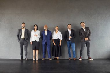 Gri stüdyo duvarında izole edilmiş, resmi giyinmiş, şirket toplantısına hazır altı çalışanın tam boy çekimi. Kapalı alanda poz veren şirket çalışanlarının tam vücut portresi..