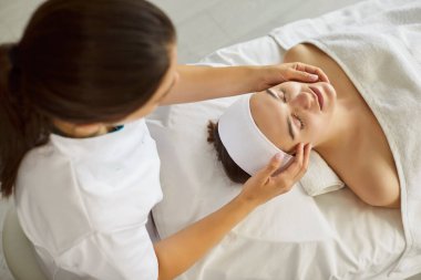 Klinikteki güzellik seansı, tedavi yatağında gözleri kapalı uzanan kadın, güzellik uzmanının elleri nazikçe yüzüne, kafasına, kaplıcadaki tapınaklara masaj yapıyor, rahatlama, kadın bakımı ve gençleşme.