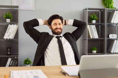 Masada oturan mutlu iş adamının ofis işinden memnun bir ifadeyle ayrılması. Patron pozunda oturan, sandalyeye yaslanan, elleri başının arkasında olan bir adam. Rahat, rahat yaşam konsepti