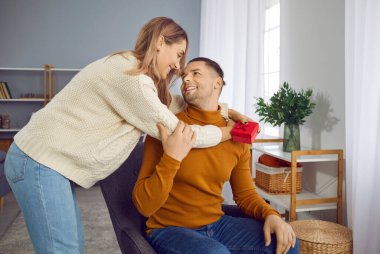 Hediye kutusuyla birbirlerine şefkatle bakan mutlu genç çift. Örgü örmüş kazaklar giyen romantik çift evde sarılıyor, güzel kız kırmızı hediye kutusu tutuyor.