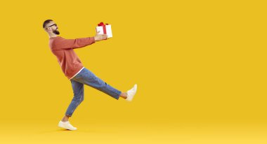 Hediye kutusu taşıyan mutlu bir adam. Sıradan kıyafetlerle gezen ve parlak turuncu boş bir fotokopi stüdyosunun arka planında yürüyen komik bir gencin yan görüntüsü. Tatil kavramı