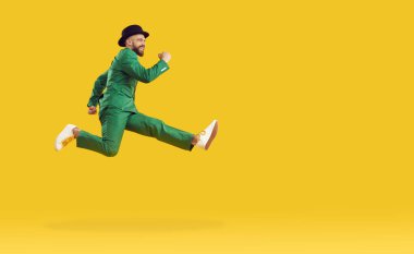 Mutlu, yeşil takım elbiseli ve siyah şapkalı genç adam St. Patrick Günü partisine koşuyor. Hızlı koşuyor ve havada yükseğe zıplıyor. Parlak sarı bir arka planda izole edilmiş.
