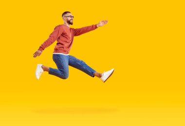 Heyecanlı pozitif adam hedeflerine ulaşmak için hızlı bir şekilde koşup zıplıyor. Uzun ve mutlu bir adam, günlük giysiler içinde havada süzülüyor turuncu arka planda adım atıyor. Profilde yan görünüm.
