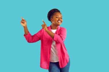 Neşeli, pozitif kıvırcık Afrikalı Amerikalı kadın turkuaz arka planda fotokopi alanında dans ediyor. Pembe gömlek, kot pantolon ve beyaz tişört giyiyor, gülümsüyor ve kameraya bakıyor..