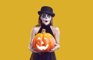 Cadılar Bayramı kostümlü çocuk. Şapkalı, şeker kafalı kız sarı arka planda tek başına duruyor, balkabağı Jack 'i tutuyor ve komik, şaşırmış yüz ifadesiyle kameraya bakıyor.