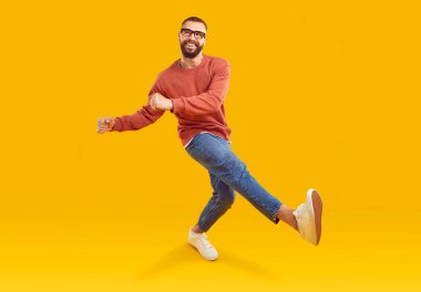 Sıradan giyinen mutlu adam dans ediyor ve eğleniyor. Sarı arka planda, rahat turuncu kazaklı, kot pantolonlu, spor ayakkabılı ve gözlüklü neşeli ve çekici bir genç. Moda konsepti