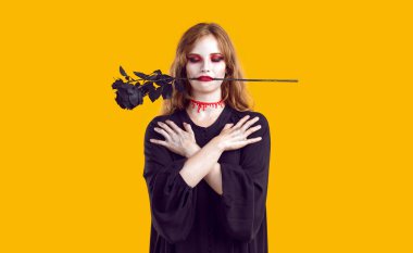 Ürkütücü Cadılar Bayramı kostümü giymiş genç bir kadının stüdyo fotoğrafı. Gözleri kapalı, boynu kan içinde, ağzı siyah bir gül içinde sarı arka planda tek başına duran ve göğsünde çapraz el ele tutuşan ölü bir kız.