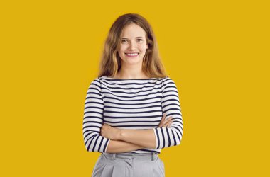 Sıradan giyinmiş mutlu bir kadının stüdyo portresi. Sarışın, uzun saçlı, çizgili bluzlu, kolları çapraz duran, turuncu sarı arka planda izole edilmiş, kameraya bakıp gülümseyen bir kız.