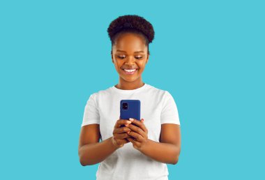 Beyaz kıvırcık saçlı, beyaz tişörtlü mutlu bir Afro-Amerikan kadını akıllı telefon ekranına bakıyor, mesaj okuyor ya da video izliyor, turkuaz arka planda görüntülü sohbet yapıyor..