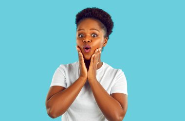 Şaşırmış kadının stüdyo portresi. Katı mavi arka planda izole edilmiş güzel Afro-Amerikan bir kızın fotoğrafı. Tuhaf, şaşkın, mutlu yüz ifadesi olan bir şeye bakıyor.