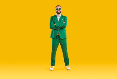 Yeşil takım elbiseli ve güneş gözlüklü neşeli ve neşeli bir adamın portresi. Aziz Patrick Günü 'nü kutlayan komik adam elleri bağlı turuncu arka planda kameraya gülümsüyor..