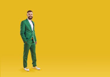 Kutlama tarzı. Portakal rengi arka planda izole edilmiş modern takım elbiseli neşeli, havalı ve şık bir adamın portresi. Yeşil takım elbiseli yakışıklı sakallı adam fotokopi alanının yanında duruyor. Aziz Patrik