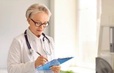 Not panosuyla pratisyen hekim. Beyaz önlüklü, gözlüklü, kontrol listesi yazan orta yaşlı olgun bir kadın ya da genel muayene sonucu, tıbbi belge dolduran ya da tedavi öneren. Tıp konsepti