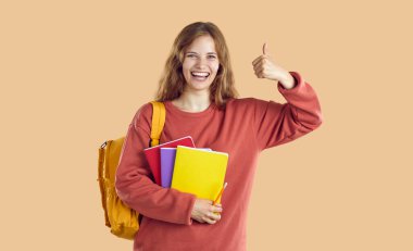 Neşeli, komik bayan öğrenci açık bej arka planda onay işareti ya da onay jesti gösteriyor. Sırt çantalı ve ders kitaplı neşeli Avrupalı kız baş parmağını gösteriyor. Eğitim konsepti. Web pankartı.
