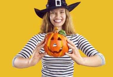 Cadılar Bayramı tarzında neşeli bir kadının ellerinde oyulmuş komik suratlı küçük dekoratif balkabağı. Cadıların şapkasıyla turuncu arka planda gülümseyen kız. Cadılar Bayramı konsepti Seçici odak.