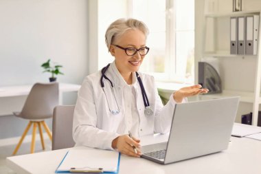 Dost canlısı kıdemli kadın doktor, video bağlantısı yoluyla hastaya çevrimiçi danışmanlık yaparken modern teknolojileri geliştiriyor. Doktor ofisteki iş yerinde oturuyor. Teletıp kavramı.