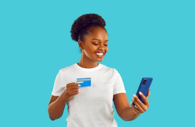 Elinde cep telefonu tutarken kredi kartı gösteren Afro-Amerikalı bir kadının portresi, mavi arka planda izole edilmiş. Kablosuz satın alma çevrimiçi mağaza konsepti.