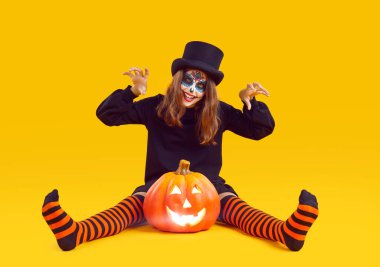 Ürkütücü kostümlü çocuk portresi. Küçük çocuk Cadılar Bayramı 'nda eğleniyor. Kafatası makyajlı, silindir şapkalı ve çizgili turuncu siyah çoraplı kız sarı stüdyo arka planında balkabağıyla yerde oturuyor.