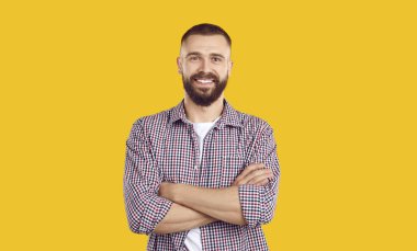 Kolları bağlı genç bir adamın stüdyo portresi. Mutlu, yakışıklı, sakallı, sıradan bir gömlek giyen ve sarı arka planda tek başına duran genç adam kameraya bakıyor ve gülümsüyor.