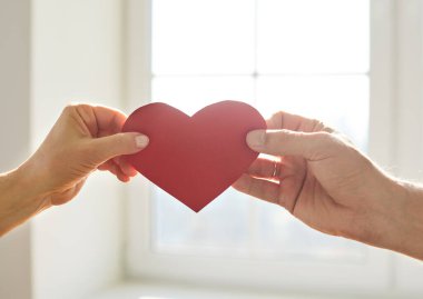 Kadın ve erkeğin elleri, kırmızı kağıt kalbi ellerinde tutuyorlar. Arka planda, evde duruyorlar. Sevgi ve özen göstergesi olarak kalbi olan bir çifti yakınlaştırın. Romantik ilişkiler konsepti.