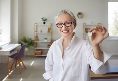 Yeni bir evde yeni bir hayata başlamak. Kapalı alanda gülümseyen güzel, bekar, olgun, beyaz kadın, iş kadını, emlakçı ya da emlakçı, oturma odasında duruyor ve evin anahtarını gösteriyor.