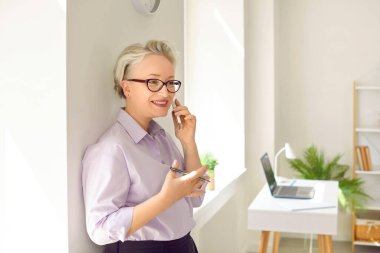 Ofisteki arkadaş canlısı kıdemli iş kadını potansiyel müşteriyle cep telefonuyla konuşuyor. Güzel ve şık beyaz saçlı, akıllı telefonlu kadın iş telefonuna gülümseyerek cevap veriyor..