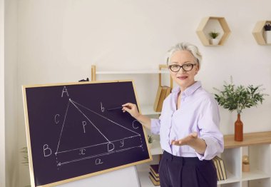 Okul sınıfında ders veren profesyonel bir matematik öğretmeninin portresi. Gözlüklü mutlu, olgun bir kadın tebeşir tahtasını işaret ediyor ve geometri teoremi hakkında bir soru soruyor.