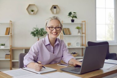 Tişörtlü, gözlüklü, kısa beyaz saçlı, masa başında oturan, dizüstü bilgisayarı kullanan, not defterine yazan, kameraya bakıp gülümseyen mutlu, zarif bir son sınıf öğrencisiydi.