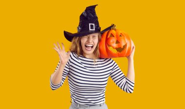 Komik cadı kız çığlık atıyor. Omuzunda turuncu bir balkabağı olan, sarı arka planda izole bir şekilde duran ve yüksek sesle bağıran cadı şapkalı ürkek genç kadın. Cadılar Bayramı, korku konsepti