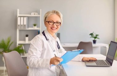 Deneyimli, gri saçlı, gülümseyen kadın doktor hastaya internet üzerinden danıştıktan sonra kameraya döner ve hastane ofisinde dizüstü bilgisayarla masada oturur.