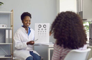 Mutlu, güzel, koyu tenli, genç doktor kadın göz doktoru kıvırcık saçlı küçük kızın görüşünü kontrol etmek için semboller gösteriyor. Sağlık ve sağlıklı yaşam tarzı kavramı.