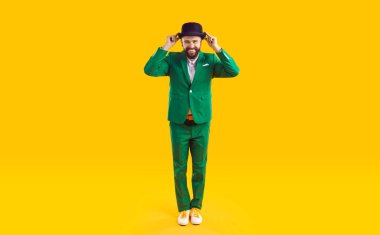 Stüdyonun sarı arka planında duran yeşil takım elbiseli ve siyah şapkalı mutlu, neşeli, neşeli, komik, gülümseyen, sakallı adamın tüm vücut çekimi. Moda ve Aziz Patrick Günü konsepti