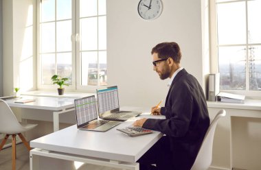 Erkek muhasebeci iki modern dizüstü bilgisayar üzerinde çalışıyor. Genç adam ofis masasında oturuyor, evrak işleriyle uğraşıyor, bilgisayar ekranlarına bakıyor, iş sayfalarıyla uğraşıyor, verileri kontrol ediyor ve notlar alıyor.