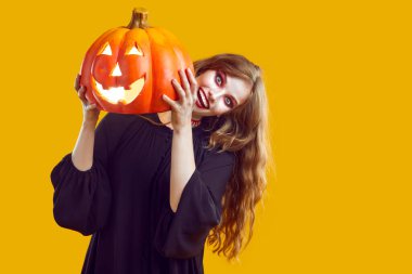 Korkunç makyajlı, dalgalı açık renk saçlı, siyah cüppeli, elinde balkabağı olan deli bir kadın ellerinde sarı arka plan ve fotokopi alanı var. Cadılar Bayramı afişi, kostüm, kutlama.