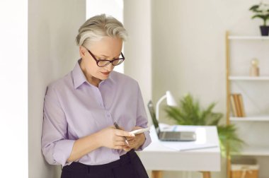 Modern iş kadını cep telefonu kullanıyor. Ofis işyerindeki pencerede duran, elinde kalem ve cep telefonu tutan, ihbarnameleri okuyan ya da iş notları arayan üst düzey üst düzey kadın.
