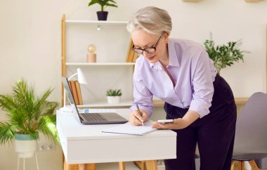 Kıdemli iş kadını cep telefonu ekranına bakarken not defterine not alıyor. Başarılı stil sahibi, olgun bir kadın. Parlak modern ofisinde çalışıyor. İş ve yaşlılar konsepti.
