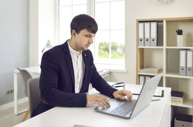 Erkek ofis çalışanı ofiste otururken elektronik belgelerle dizüstü bilgisayarda çalışıyor. Klavyede yazan, e-posta gönderen ya da hesap tablolarını dolduran ceketli zeki bir adam. İş konsepti.