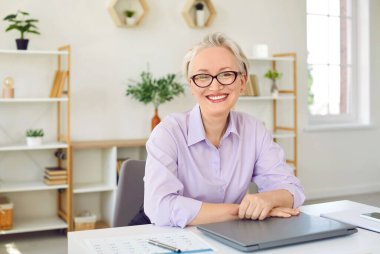 Mutlu, gri saçlı, beyaz, yaşlı, beyaz iş kadını ev ofisinin masasında otururken uzaktan iş yaptıktan sonra elleri kapalı kameraya bakıp gülümsüyor. Serbest çalışma, girişimcilik.