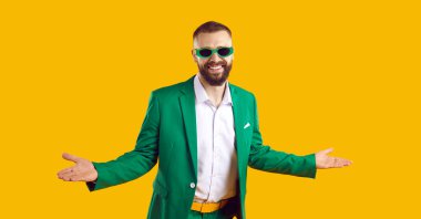 Yeşil takım elbiseli ve güneş gözlüklü komik sakallı genç adam sarı arka planda tek başına duruyor, kollarını açıyor, gülümsüyor ve Aziz Patrick Günü partisine hoş geldiniz diyor. Moda ve parti konsepti