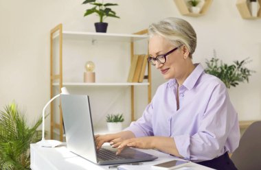 Bilgisayarda çalışan bir iş kadını. Tişörtlü ve gözlüklü mutlu orta yaşlı iş kadını rahat ofisinde oturuyor ve dizüstü bilgisayarında daktilo yazıyor.