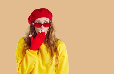 Şirin, şok olmuş genç bir kadın ilginç bir sırrı eliyle ağzını kapatıyor. Modaya uygun bereli, gözlüklü, eldivenli ve sweatshirt 'lü beyaz kız bej arka planda başka tarafa bakarken.