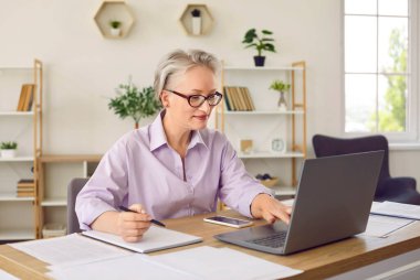 Üst düzey iş kadını, ofisinde çalışırken elektronik ve kağıt belgelerini karşılaştırıyor. Güzel, olgun bir kadın masada oturuyor, dizüstü bilgisayarda yazıyor ve kağıt belgelere notlar yazıyor..