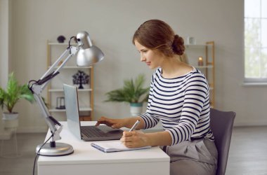 Ev bilgisayarında çalışan ve not defterine çeşitli notlar alan yaratıcı serbest çalışan kadın. Masada oturan, uzaktan çalışan, video izleyen ya da internette çalışan genç bir kadının yan görüntüsü.