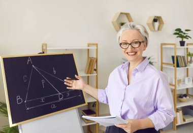 Okulunda neşeli bir matematik öğretmeninin portresi. Tişörtlü ve gözlüklü mutlu orta yaşlı kadın tebeşir tahtasına geometri teoremi ile gülümsüyor ve işaret ediyor.
