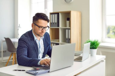 Ofiste dizüstü bilgisayar üzerinde çalışırken finansal piyasa araştırması yapan konsantre erkek yönetici. Takım elbiseli ve gözlüklü ciddi bir adam masada oturuyor ve dizüstü bilgisayar ve hesap makinesiyle çalışıyor..