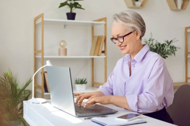 Ev ofisinden uzaktan çalışan mutlu, şık, olgun yaşlı kadın dizüstü bilgisayarda yazıyor. 60 'ların gülümseyen iş kadını uzaktan kumanda olarak bilgisayar kullanıyor, evinde oturuyor..